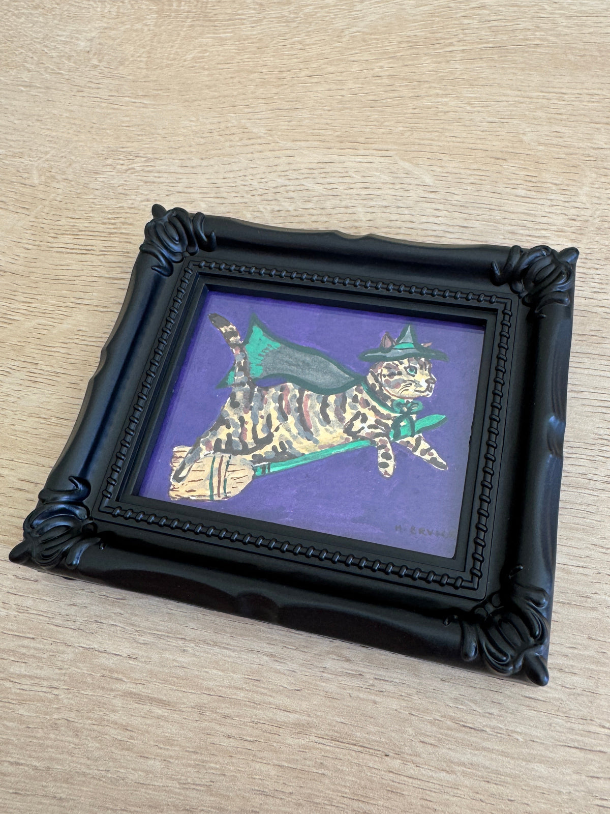 A small framed art print featuring a whimsical illustration of a striped cat dressed as a witch on a broomstick against a purple background, housed in an ornate black frame, placed on a light wood surface.